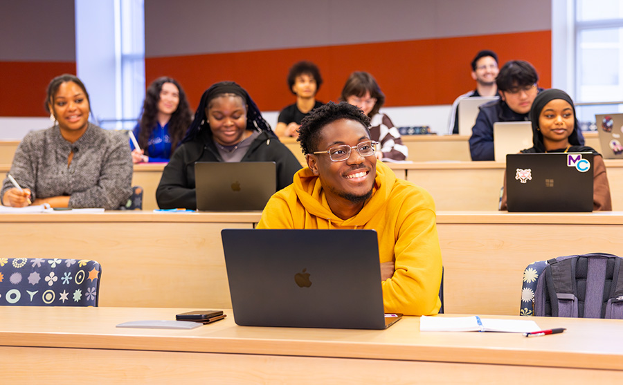 Patrick in a yellow sweater smiles at his laptop, seated in a classroom with other students behind him.